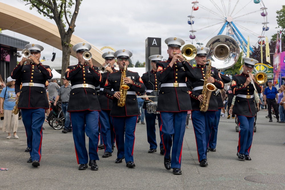 A United States Marine Corps marching band performs at what appears to be a state fair or outdoor public event. The musicians are dressed in formal dress blues uniforms and are playing various brass and woodwind instruments while marching through a crowded fairground. A Ferris wheel and carnival attractions are visible in the background.