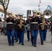 A United States Marine Corps marching band performs at what appears to be a state fair or outdoor public event. The musicians are dressed in formal dress blues uniforms and are playing various brass and woodwind instruments while marching through a crowded fairground. A Ferris wheel and carnival attractions are visible in the background.