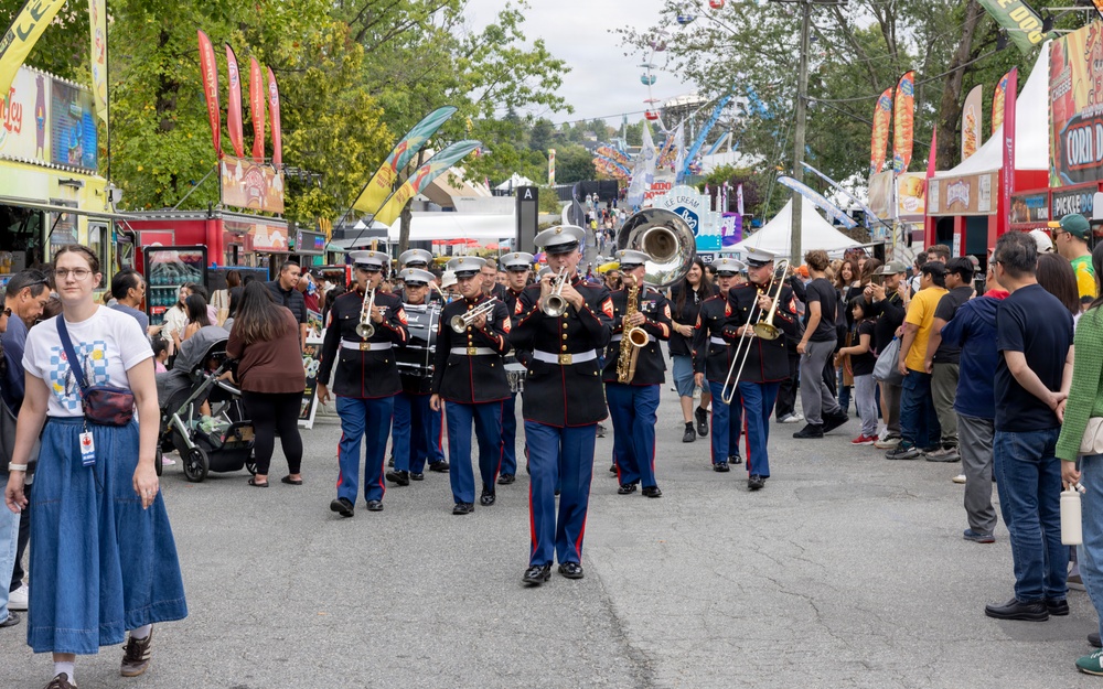 A U.S. Marine Corps marching band performs during a parade at what appears to be a large outdoor fair or festival. The uniformed musicians are playing brass and wind instruments while marching down a crowded midway lined with food vendors and colorful banners. Spectators line both sides of the path watching the performance.
