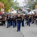 A U.S. Marine Corps marching band performs during a parade at what appears to be a large outdoor fair or festival. The uniformed musicians are playing brass and wind instruments while marching down a crowded midway lined with food vendors and colorful banners. Spectators line both sides of the path watching the performance.