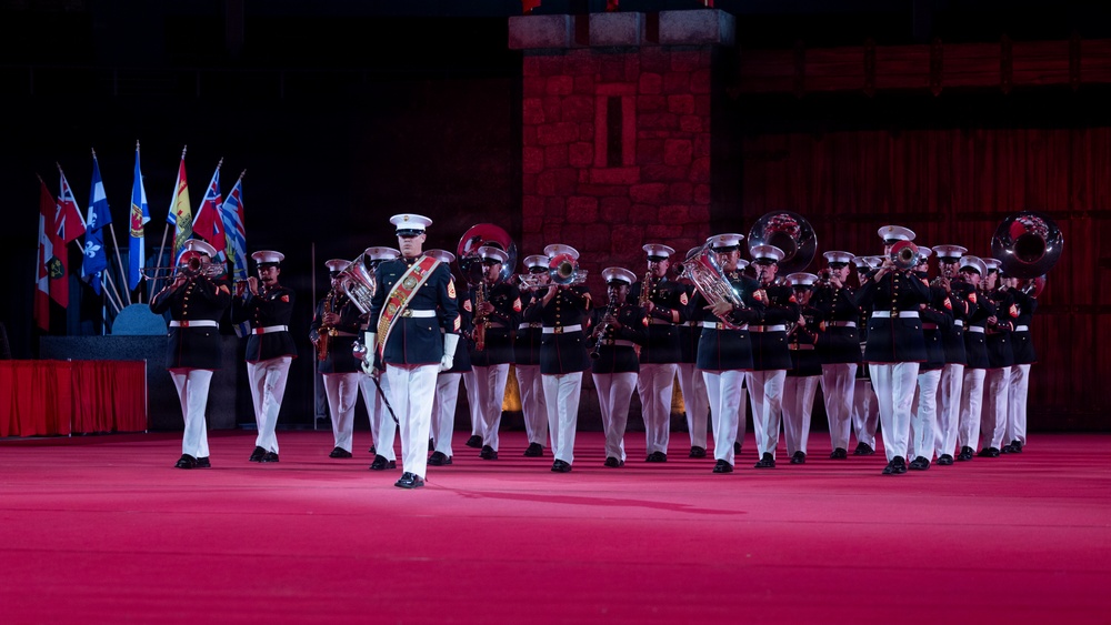 A military marching band performs on a red-lit stage or parade ground, wearing dark dress uniforms with white trousers and white peaked caps. The ensemble includes brass instruments such as tubas, trombones, and trumpets, led by a drum major or band leader in the foreground. Multiple national flags are visible in the background, suggesting an international military tattoo or ceremonial event.