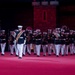 A military marching band performs on a red-lit stage or parade ground, wearing dark dress uniforms with white trousers and white peaked caps. The ensemble includes brass instruments such as tubas, trombones, and trumpets, led by a drum major or band leader in the foreground. Multiple national flags are visible in the background, suggesting an international military tattoo or ceremonial event.