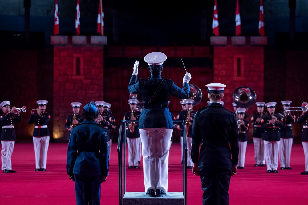 A military band performance is taking place in a ceremonial indoor venue with red lighting and castle-like stone architecture. A conductor in a dark blue uniform with white trousers stands on a podium, baton raised, directing a large ensemble of musicians in white and red uniforms. Canadian flags are visible in the background, suggesting this is a Canadian military tattoo or ceremonial event.