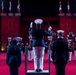 A military band performance is taking place in a ceremonial indoor venue with red lighting and castle-like stone architecture. A conductor in a dark blue uniform with white trousers stands on a podium, baton raised, directing a large ensemble of musicians in white and red uniforms. Canadian flags are visible in the background, suggesting this is a Canadian military tattoo or ceremonial event.