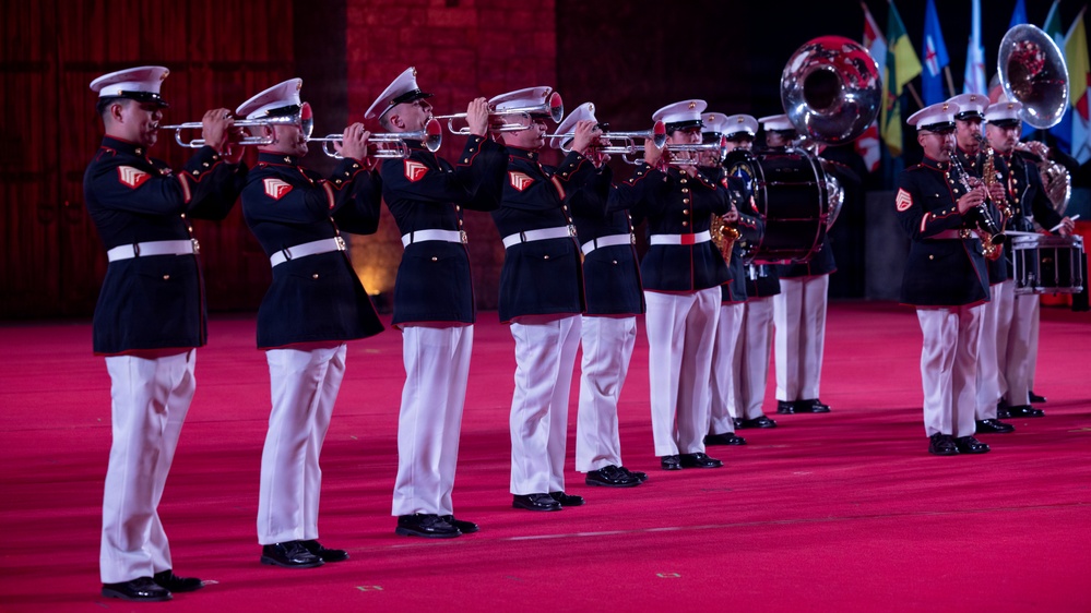 A military marching band performs on a red-carpeted stage or parade ground. The musicians are dressed in formal dark navy dress uniforms with white trousers, white caps, and white belts, consistent with U.S. Marine Corps dress blues. The ensemble includes trumpet/bugle players in the foreground, with sousaphones, saxophones, and percussion visible in the background.