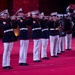 A military marching band performs on a red-carpeted stage or parade ground. The musicians are dressed in formal dark navy dress uniforms with white trousers, white caps, and white belts, consistent with U.S. Marine Corps dress blues. The ensemble includes trumpet/bugle players in the foreground, with sousaphones, saxophones, and percussion visible in the background.