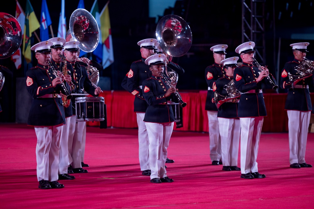 A United States Marine Corps band is performing at a formal military ceremony or tattoo event on a red-carpeted stage. The musicians are dressed in full dress blues with white covers (caps) and white trousers, playing a variety of instruments. Multiple national or organizational flags are visible in the background, suggesting an international or ceremonial event.