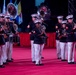 A United States Marine Corps band is performing at a formal military ceremony or tattoo event on a red-carpeted stage. The musicians are dressed in full dress blues with white covers (caps) and white trousers, playing a variety of instruments. Multiple national or organizational flags are visible in the background, suggesting an international or ceremonial event.