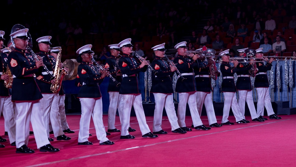A U.S. Marine Corps marching band is performing on a red-carpeted indoor arena floor. The musicians are dressed in formal dress blues uniforms with white trousers and white peaked caps, playing a variety of wind instruments. An audience is visible in the background seated in stadium-style seating.