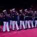A U.S. Marine Corps marching band is performing on a red-carpeted indoor arena floor. The musicians are dressed in formal dress blues uniforms with white trousers and white peaked caps, playing a variety of wind instruments. An audience is visible in the background seated in stadium-style seating.