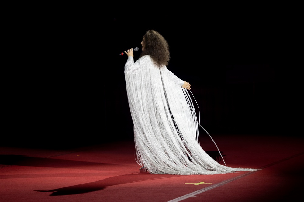 A performer stands on a red-carpeted stage wearing an elaborate white fringe gown that cascades to the floor. The figure is photographed from behind, holding a microphone and appearing to sing, set against a completely dark background. The dramatic costume and staging suggest a high-profile live performance or ceremony.