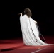 A performer stands on a red-carpeted stage wearing an elaborate white fringe gown that cascades to the floor. The figure is photographed from behind, holding a microphone and appearing to sing, set against a completely dark background. The dramatic costume and staging suggest a high-profile live performance or ceremony.