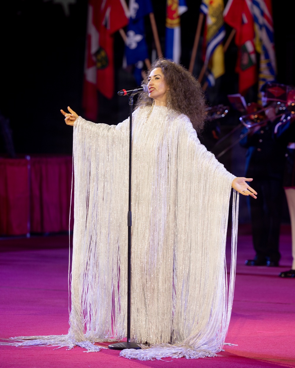 A female performer is singing at a microphone on a stage, wearing an elaborate white floor-length gown with extensive fringe detailing. She is performing at what appears to be a formal or national event, evidenced by multiple flags visible in the background. Her arms are outstretched in an expressive performance gesture.