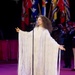 A female performer is singing at a microphone on a stage, wearing an elaborate white floor-length gown with extensive fringe detailing. She is performing at what appears to be a formal or national event, evidenced by multiple flags visible in the background. Her arms are outstretched in an expressive performance gesture.