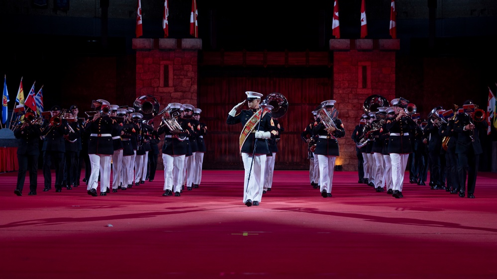 A large military band performs on a red-lit indoor arena floor, with a central figure in dress uniform executing a salute. The performers wear dark navy/black uniforms with white trousers and white peaked caps, carrying brass instruments including tubas and trumpets. A castle-like stone backdrop is decorated with Canadian flags, suggesting this is a Canadian military tattoo or ceremonial performance.
