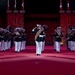 A large military band performs on a red-lit indoor arena floor, with a central figure in dress uniform executing a salute. The performers wear dark navy/black uniforms with white trousers and white peaked caps, carrying brass instruments including tubas and trumpets. A castle-like stone backdrop is decorated with Canadian flags, suggesting this is a Canadian military tattoo or ceremonial performance.