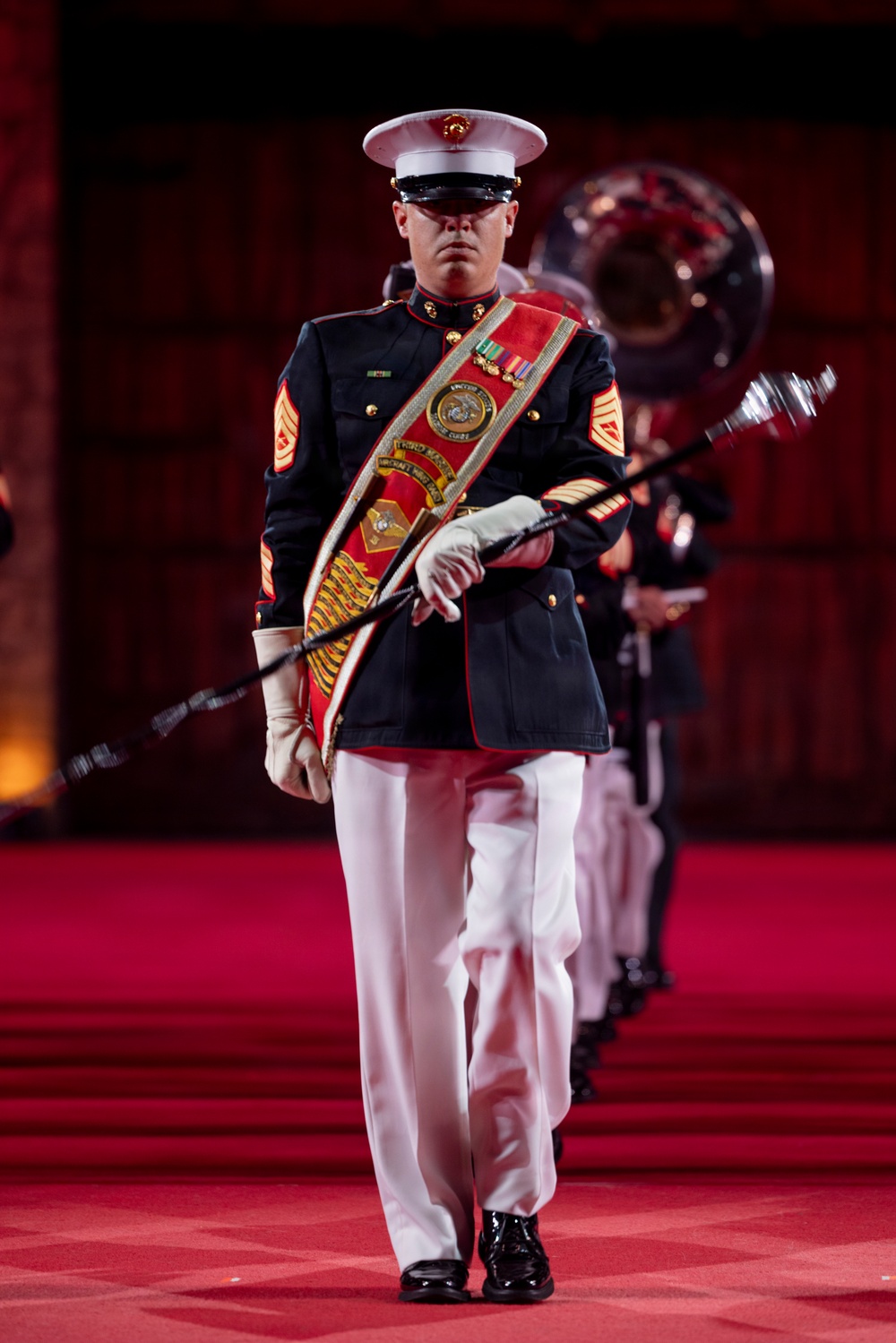 A United States Marine Corps drum major leads a marching band in formal dress blues uniform during what appears to be a ceremonial performance. The lead figure wears a decorative red sash/baldric with the USMC Eagle, Globe, and Anchor emblem and carries a mace baton. The formation marches on a red-carpeted stage or performance floor in an indoor venue.