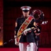 A United States Marine Corps drum major leads a marching band in formal dress blues uniform during what appears to be a ceremonial performance. The lead figure wears a decorative red sash/baldric with the USMC Eagle, Globe, and Anchor emblem and carries a mace baton. The formation marches on a red-carpeted stage or performance floor in an indoor venue.