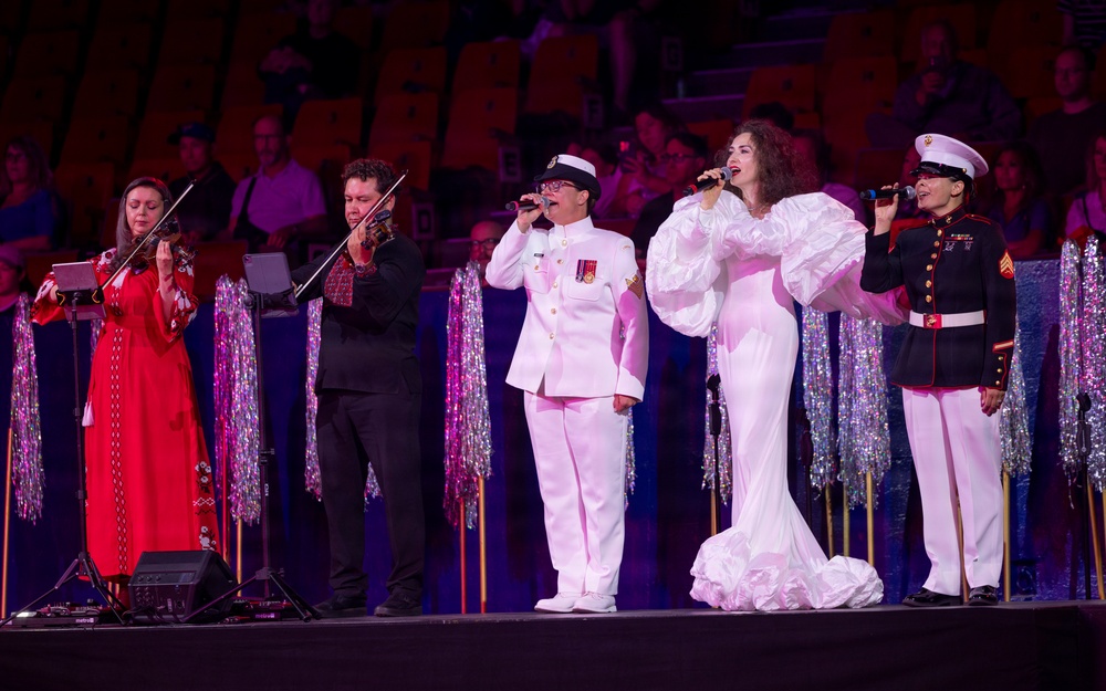 A live performance on a stage featuring five performers, including two military personnel in uniform, a civilian woman in an elaborate white gown, and two musicians playing violins. The event appears to be a formal or ceremonial concert held in a large venue with an audience visible in the background. The stage is decorated with silver tinsel curtains and gold poles.