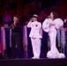 A live performance on a stage featuring five performers, including two military personnel in uniform, a civilian woman in an elaborate white gown, and two musicians playing violins. The event appears to be a formal or ceremonial concert held in a large venue with an audience visible in the background. The stage is decorated with silver tinsel curtains and gold poles.