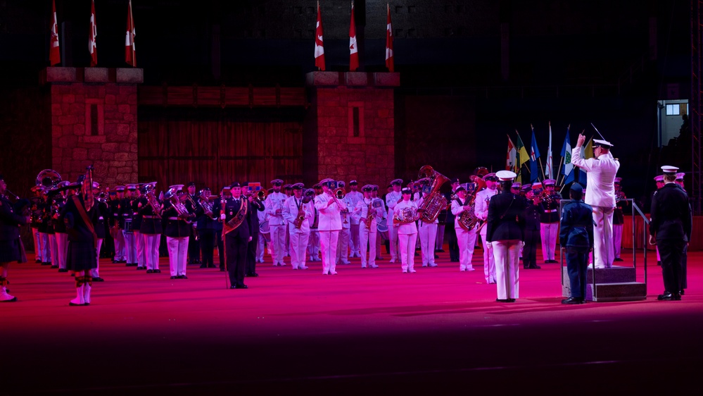 A large military band performance is taking place in an outdoor ceremonial setting, illuminated by dramatic pink/red stage lighting. Multiple uniformed musicians from what appears to be naval and military branches are assembled, with a conductor on a raised podium directing the ensemble. Canadian flags and a castle-like stone backdrop are visible, suggesting this may be a Canadian military tattoo or ceremonial parade.