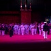 A large military band performance is taking place in an outdoor ceremonial setting, illuminated by dramatic pink/red stage lighting. Multiple uniformed musicians from what appears to be naval and military branches are assembled, with a conductor on a raised podium directing the ensemble. Canadian flags and a castle-like stone backdrop are visible, suggesting this may be a Canadian military tattoo or ceremonial parade.