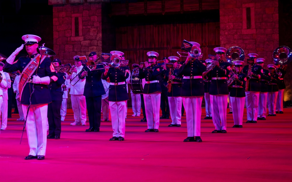A military marching band performs at a formal evening ceremony, illuminated by pink and red stage lighting. The musicians, dressed in dark navy dress uniforms with white trousers and white peaked caps, are playing brass instruments in formation. A senior officer or drum major in the foreground is saluting, wearing a distinctive red sash.