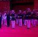 A military marching band performs at a formal evening ceremony, illuminated by pink and red stage lighting. The musicians, dressed in dark navy dress uniforms with white trousers and white peaked caps, are playing brass instruments in formation. A senior officer or drum major in the foreground is saluting, wearing a distinctive red sash.