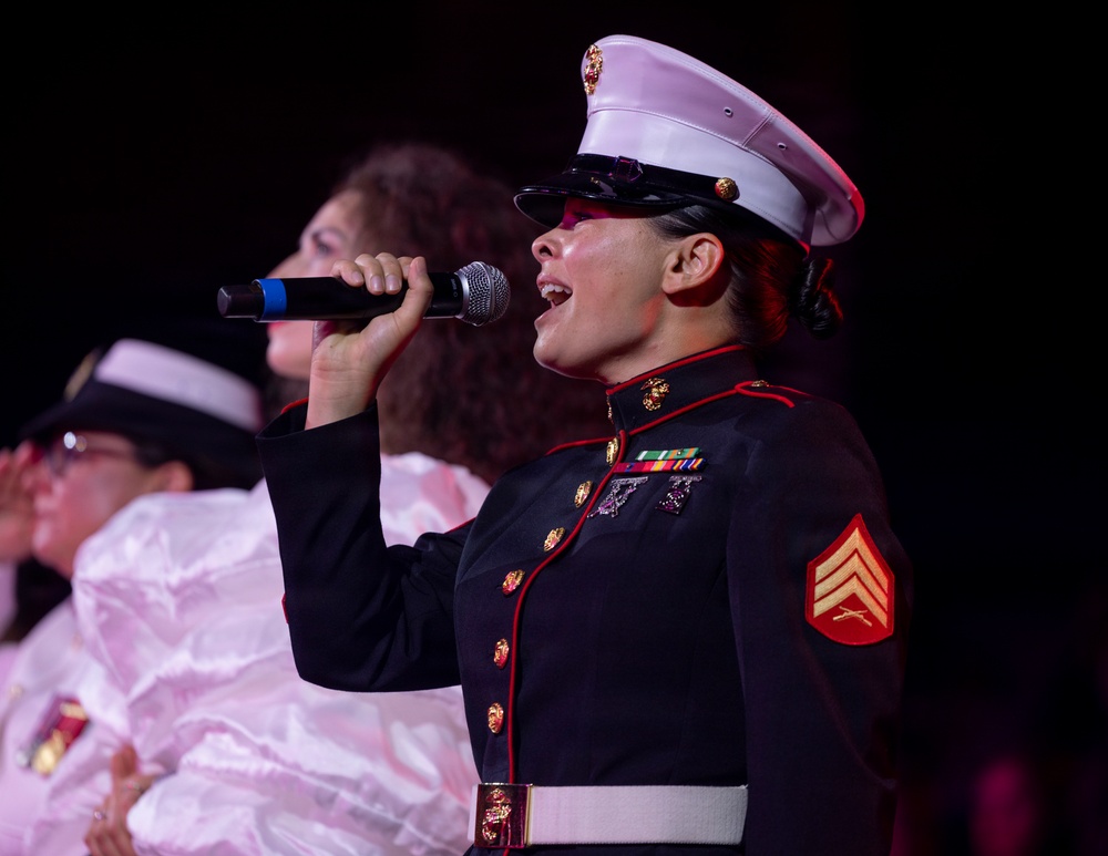 A female U.S. Marine in dress blues uniform is singing into a handheld microphone, likely performing at a formal or public event. She wears the rank insignia of a Sergeant on her sleeve and displays service ribbons on her chest. Other individuals in the background appear to be part of the same performance.