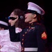 A female U.S. Marine in dress blues uniform is singing into a handheld microphone, likely performing at a formal or public event. She wears the rank insignia of a Sergeant on her sleeve and displays service ribbons on her chest. Other individuals in the background appear to be part of the same performance.