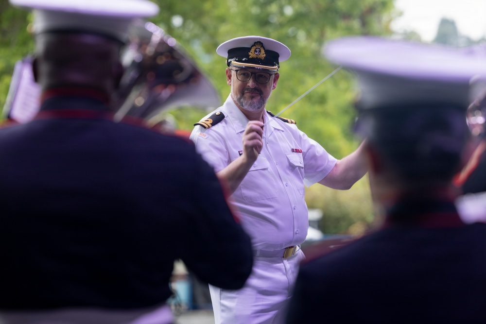 A naval officer in a white dress uniform is conducting a military band outdoors. He is holding a conductor's baton and appears to be actively directing musicians who are visible in the foreground, out of focus. The setting appears to be an outdoor ceremonial or public performance event.