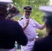 A naval officer in a white dress uniform is conducting a military band outdoors. He is holding a conductor's baton and appears to be actively directing musicians who are visible in the foreground, out of focus. The setting appears to be an outdoor ceremonial or public performance event.