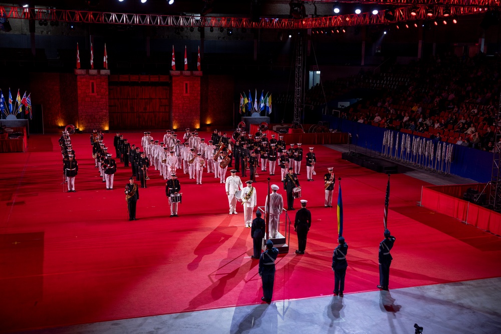 A large indoor military tattoo or ceremonial parade is taking place on a red-carpeted arena floor. Multiple military bands and honor guards in formal dress uniforms are arranged in formation, with what appears to be a central ceremony or presentation occurring at a podium. The event is held in an indoor venue with spectators in the stands, dramatic stage lighting, and decorative castle-like backdrop elements.
