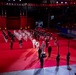 A large indoor military tattoo or ceremonial parade is taking place on a red-carpeted arena floor. Multiple military bands and honor guards in formal dress uniforms are arranged in formation, with what appears to be a central ceremony or presentation occurring at a podium. The event is held in an indoor venue with spectators in the stands, dramatic stage lighting, and decorative castle-like backdrop elements.