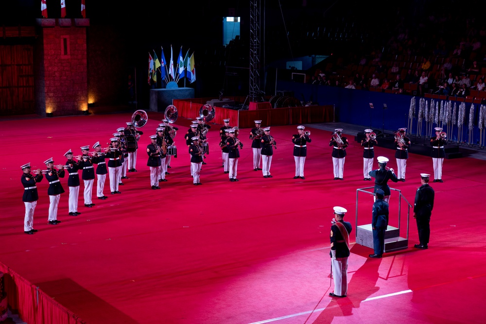 A military marching band performs on a red-carpeted indoor arena floor during what appears to be a formal military tattoo or ceremonial event. The musicians are dressed in dark dress uniforms with white trousers and white peaked caps, playing various brass and percussion instruments. A conductor stands on a podium at the front while an audience watches from the stands in the background.