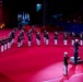 A military marching band performs on a red-carpeted indoor arena floor during what appears to be a formal military tattoo or ceremonial event. The musicians are dressed in dark dress uniforms with white trousers and white peaked caps, playing various brass and percussion instruments. A conductor stands on a podium at the front while an audience watches from the stands in the background.
