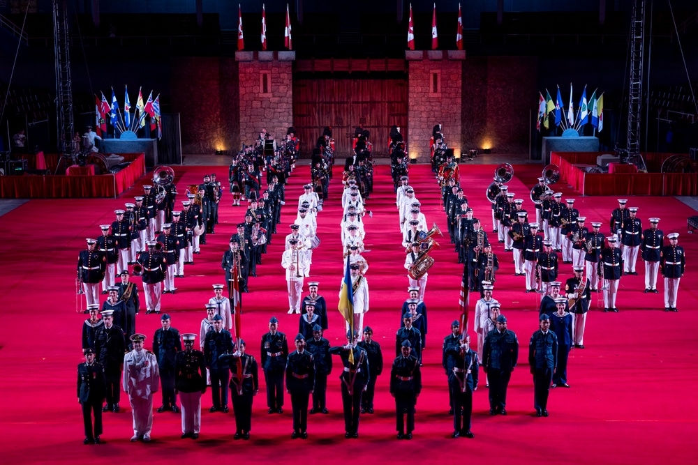 A large-scale military tattoo or ceremonial parade is taking place on a red-carpeted indoor arena floor. Multiple military bands and honor guards from various nations are assembled in formal formation, with a castle-like backdrop and national flags displayed. The event appears to be an international military music festival or tattoo ceremony.