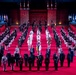 A large-scale military tattoo or ceremonial parade is taking place on a red-carpeted indoor arena floor. Multiple military bands and honor guards from various nations are assembled in formal formation, with a castle-like backdrop and national flags displayed. The event appears to be an international military music festival or tattoo ceremony.