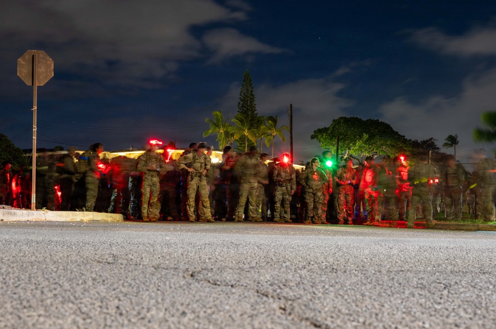 A nighttime photograph showing a large group of military personnel in camouflage uniforms gathered along a roadside. The image was taken with a long exposure, causing motion blur among the figures. Multiple red and green lights are visible among the group, likely from tactical light devices or safety markers.