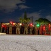 A nighttime photograph showing a large group of military personnel in camouflage uniforms gathered along a roadside. The image was taken with a long exposure, causing motion blur among the figures. Multiple red and green lights are visible among the group, likely from tactical light devices or safety markers.