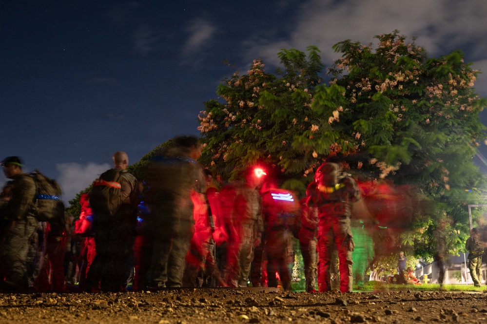 A nighttime outdoor scene showing a group of individuals in military or tactical gear illuminated by red, blue, and green lights. The long-exposure photograph creates motion blur across the figures, suggesting movement or activity during a nighttime operation or exercise. Flowering trees are visible in the background against a deep blue night sky.