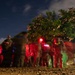 A nighttime outdoor scene showing a group of individuals in military or tactical gear illuminated by red, blue, and green lights. The long-exposure photograph creates motion blur across the figures, suggesting movement or activity during a nighttime operation or exercise. Flowering trees are visible in the background against a deep blue night sky.