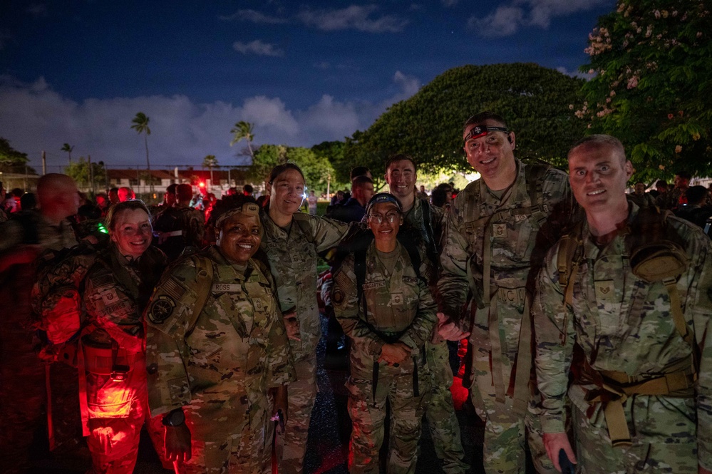 A group of military personnel in OCP (Operational Camouflage Pattern) uniforms pose together for a photo at night, likely during a ruck march or memorial event. The setting appears to be an outdoor gathering area with tropical vegetation, suggesting a location such as Hawaii. Red lights and headlamps are visible, indicating a nighttime military event with light discipline measures in place.