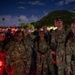A group of military personnel in OCP (Operational Camouflage Pattern) uniforms pose together for a photo at night, likely during a ruck march or memorial event. The setting appears to be an outdoor gathering area with tropical vegetation, suggesting a location such as Hawaii. Red lights and headlamps are visible, indicating a nighttime military event with light discipline measures in place.