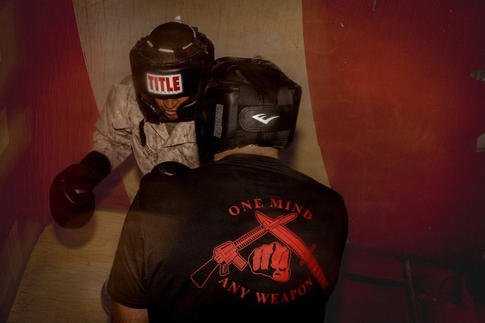 Two individuals are engaged in a close-quarters boxing or combat training session. One person wears a 'TITLE' brand headgear and military camouflage uniform with boxing gloves, while the other wears an 'Everlast' headgear and a black t-shirt reading 'ONE MIND ANY WEAPON' with crossed rifles and a fist graphic. The image has a reddish vignette overlay suggesting a training or combat sports environment.