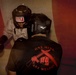 Two individuals are engaged in a close-quarters boxing or combat training session. One person wears a 'TITLE' brand headgear and military camouflage uniform with boxing gloves, while the other wears an 'Everlast' headgear and a black t-shirt reading 'ONE MIND ANY WEAPON' with crossed rifles and a fist graphic. The image has a reddish vignette overlay suggesting a training or combat sports environment.