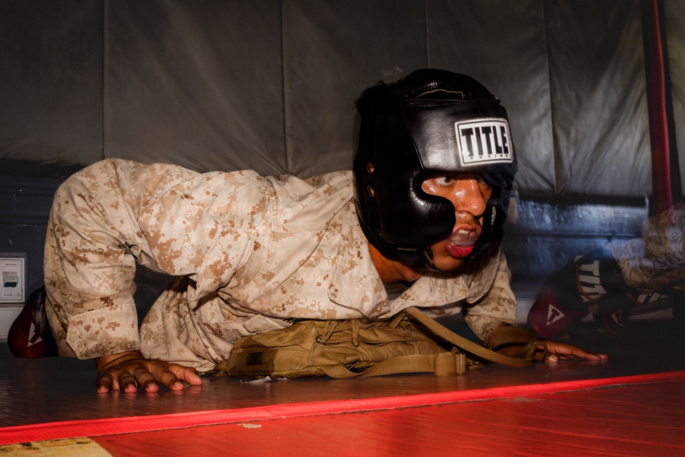 A military service member wearing USMC desert digital camouflage (MARPAT) uniform and a black 'TITLE' boxing headgear is performing push-ups or a ground exercise on a red mat. The individual appears to be sweating and exerting significant effort, suggesting an intense physical training session. The setting appears to be an indoor martial arts or combatives training facility with padded walls.