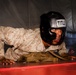 A military service member wearing USMC desert digital camouflage (MARPAT) uniform and a black 'TITLE' boxing headgear is performing push-ups or a ground exercise on a red mat. The individual appears to be sweating and exerting significant effort, suggesting an intense physical training session. The setting appears to be an indoor martial arts or combatives training facility with padded walls.