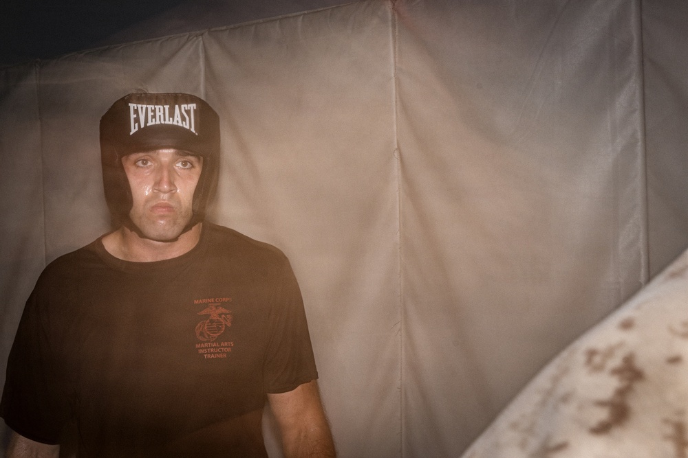 A male individual wearing an Everlast boxing headgear and a Marine Corps Martial Arts Instructor Trainer t-shirt stands against what appears to be a canvas or tent wall. His face shows perspiration, suggesting recent physical exertion. The lighting is dramatic and low, creating a moody, atmospheric composition.