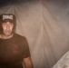 A male individual wearing an Everlast boxing headgear and a Marine Corps Martial Arts Instructor Trainer t-shirt stands against what appears to be a canvas or tent wall. His face shows perspiration, suggesting recent physical exertion. The lighting is dramatic and low, creating a moody, atmospheric composition.
